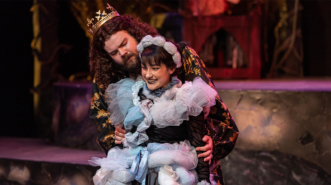 Two ornately dressed actors embrace on stage. The man’s arms are wrapped around the woman and she is smiling.