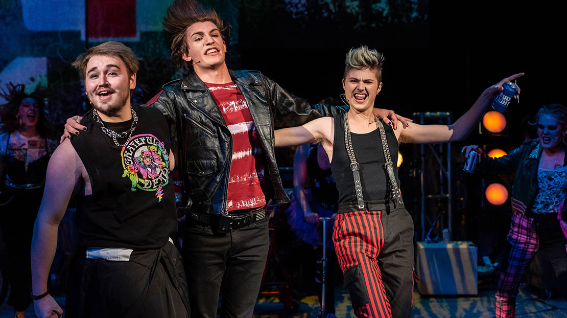 Three characters, smiling with their arms around each other’s shoulders, are dressed in red and black punk attire.